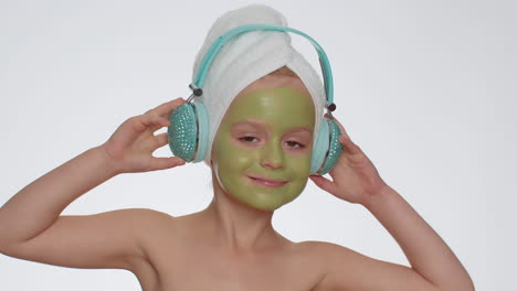 smiling young child girl moisturizing green mask on face listening to music on headphones, dancing