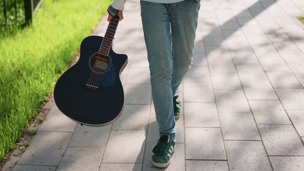 Güneş ışığıyla aydınlanmış taşlı yolda siyah gitarıyla yürüyen, kot pantolon ve spor ayakkabı giyen, akustik gitarı sapından tutan genç gitarist, rahat yürüyüş, kaldırımda benekli gölgeler, çimenli yol kenarı, indie şarkı yazarı