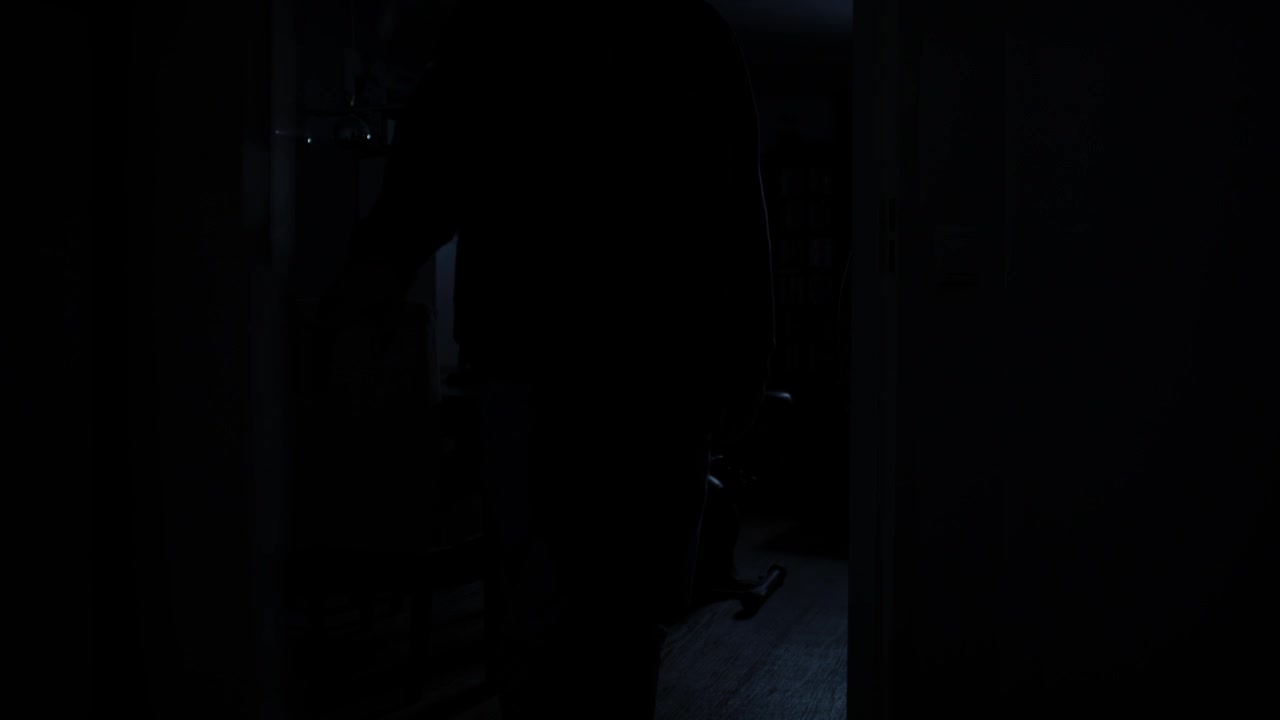 Porta di una stanza buia che si apre lentamente e rivela la silhouette di un uomo incappucciato di notte.