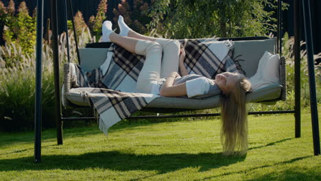 a teenage girl with long hair lies on a garden swing in the backyard of a house.