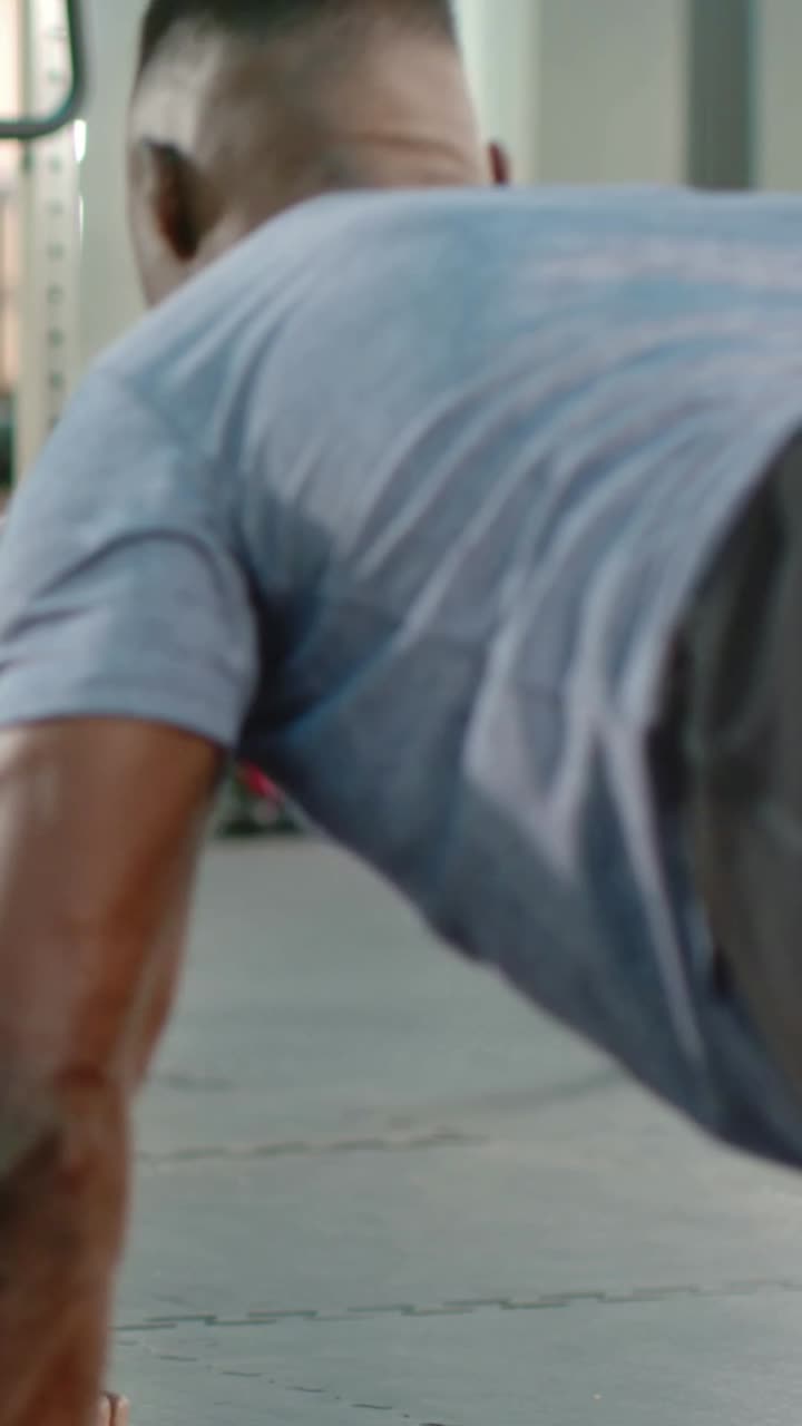 Vertical video: Lowering, pushing up, man in light-blue shirt training on gym floor, wearing watch