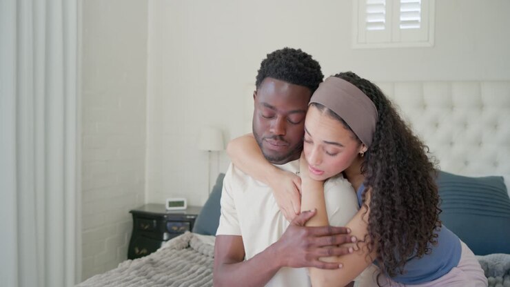 African American couple sitting on bed, woman reaching shoulder to comfort, man returning embrace