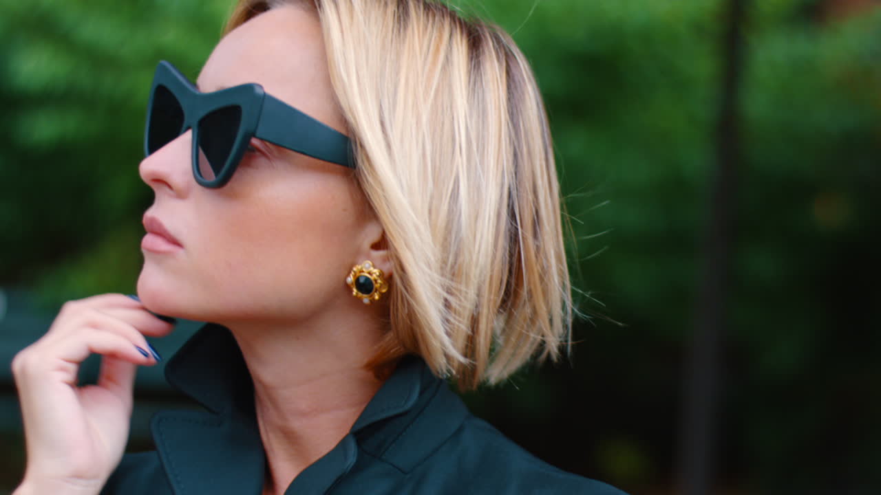 Close up of woman wearing dark geometric sunglasses. Portrait of a blonde woman wearing oversized black geometric sunglasses and a dark coat against green foliage