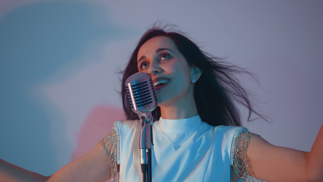 색 드레스를 입은 기쁜 가수가 빈티지 마이크를 들고 눈을 감고 손을 들고 감정을 표현합니다. 부드러운 파란색과 분홍색 빛의 배경에 설정되어 있습니다.