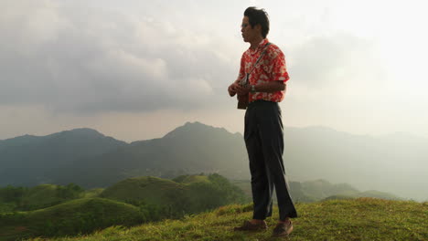 Shot-of-an-Asian-man-standing-on-top-on-a-mountain-while-playing-ukulele-on-a-cloudy-day