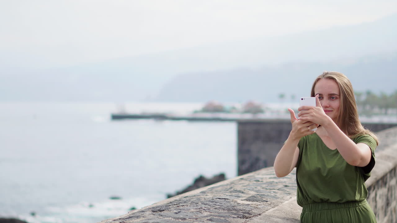 초록색 드레스를 입은 아름다운 소녀가 바다 근처의 해안가에 서서 미소 짓는 휴대전화에서 셀카를 찍습니다.