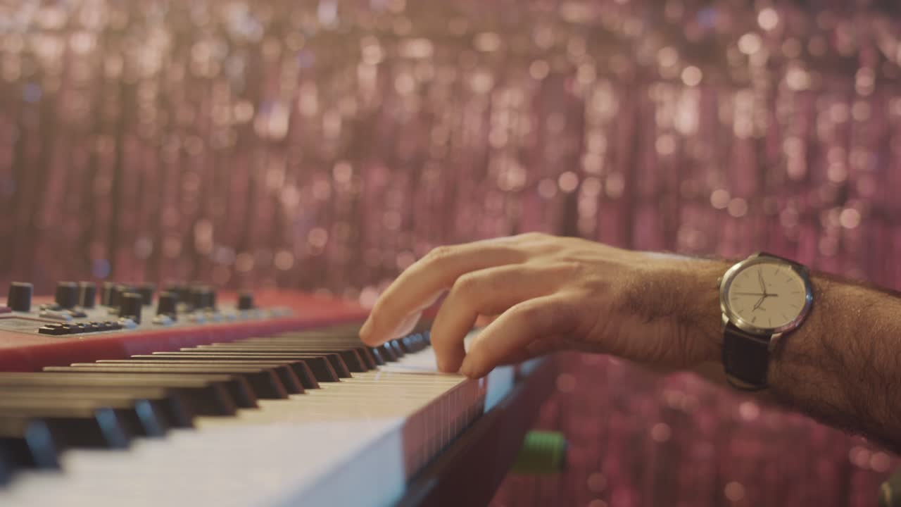A close-up shot of a musician's hands playing a melody on an electric piano. The background is a shimmering, out-of-focus curtain, creating a glamorous bokeh effect for a concert or party scene.