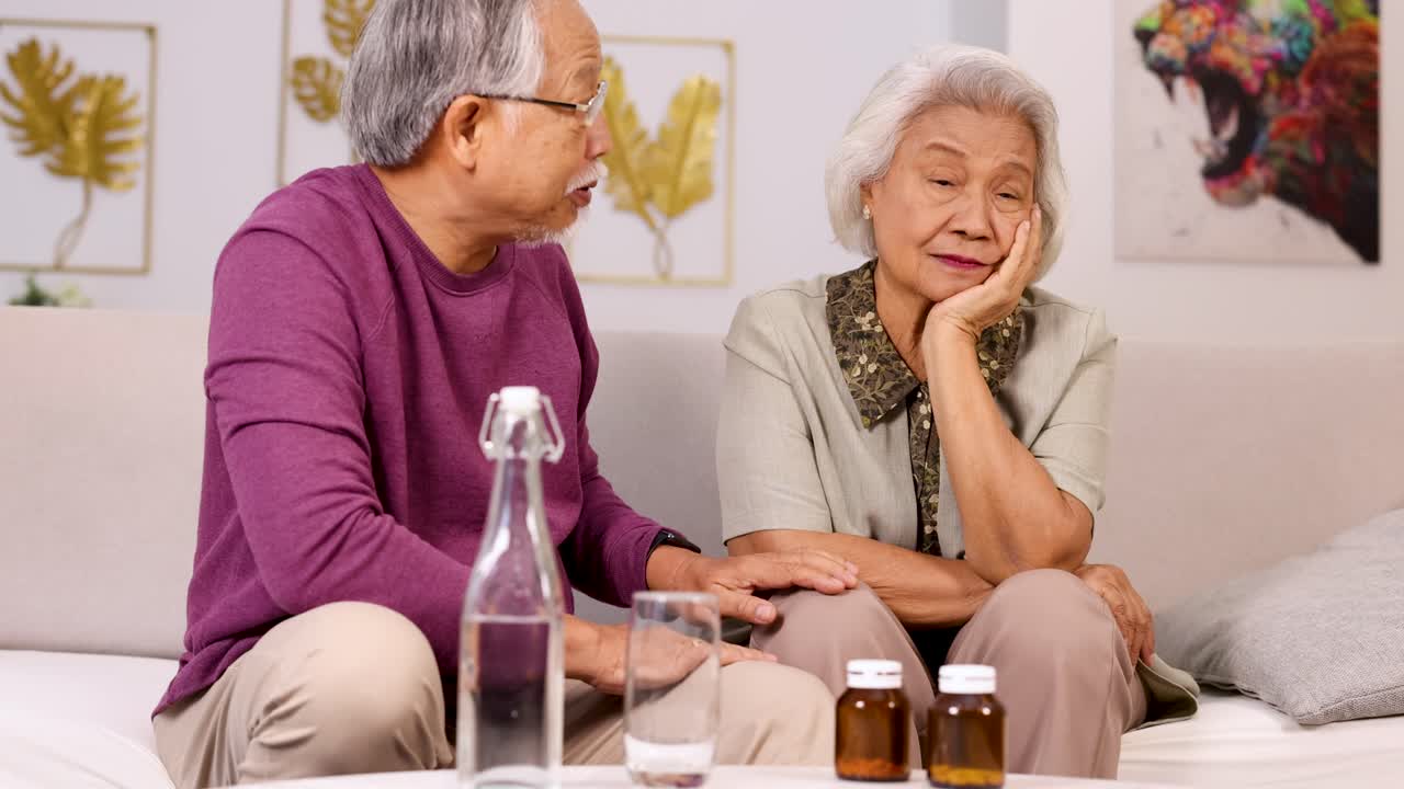 An elderly Asian couple sits on a living room sofa as the man gently checks his wife's forehead and prepares her medication under soft, natural lighting