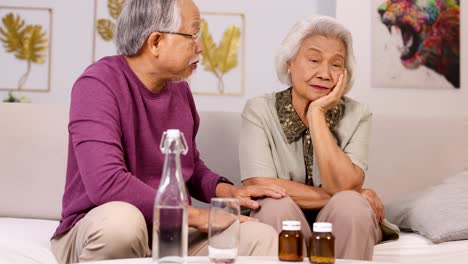 an elderly asian couple sits on a living room sofa as the man gently checks his wife's forehead and prepares her medication under soft, natural lighting