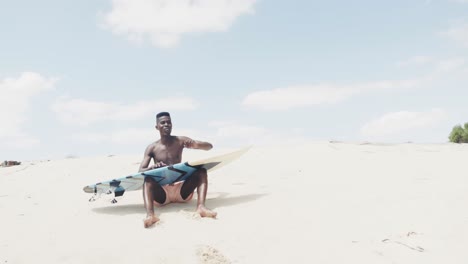 feliz hombre afroamericano con una tabla de surf, levantándose de sentarse en una playa soleada, cámara lenta