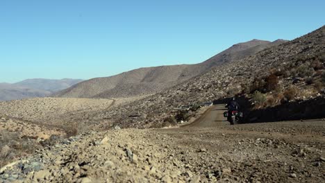 Lone-motorcyclist-rides-away-from-camera-on-remote-gravel-road,-Chile