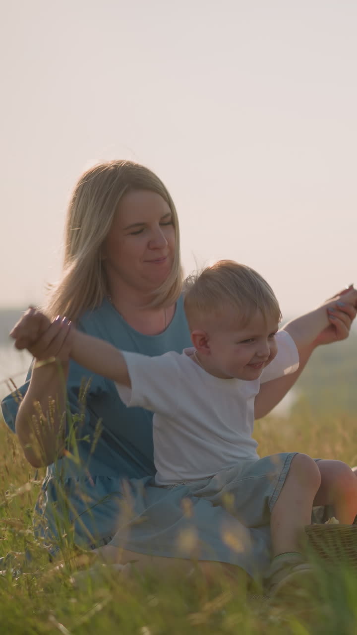 A joyful mother in a blue dress shares a tender, playful moment with her young son sitting on her lap, lifting his hands in the air. His older brother sits nearby, eating fruit from a picnic basket