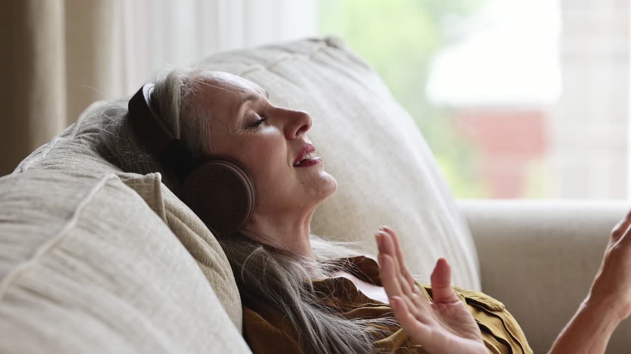 Aged female rest on couch in headphones listen to melody