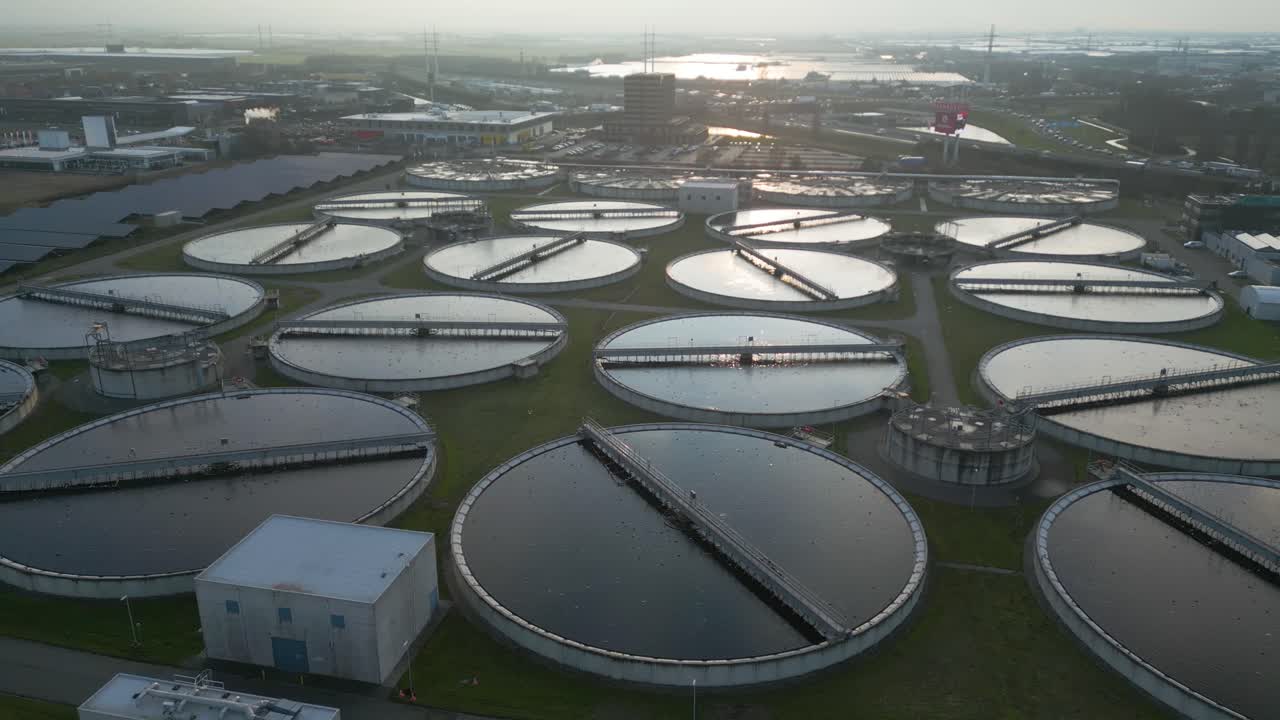 Cinematic drone approach flying over symmetrical circular wastewater clarification tanks at the Harnaschpolder treatment facility in the Netherlands during soft sunrise light