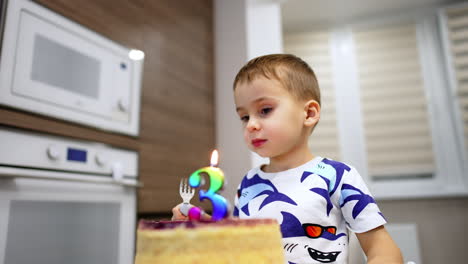 happy lovely baby boy sits looking at the cake with number three candle. kid holding a fork blows the candle and parent lights it again. low angle view.