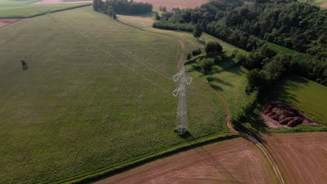 aerial of electricity tower and power lines above green farming field on a sunny summer evening, orbit drone shot