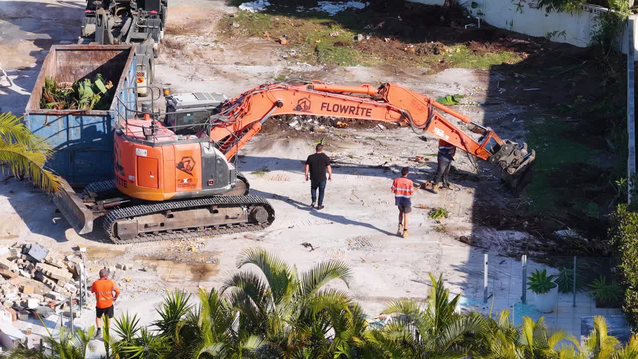 Aerial view of construction workers and an orange excavator clearing debris at a demolition site under bright daylight, with steady overhead camera angle