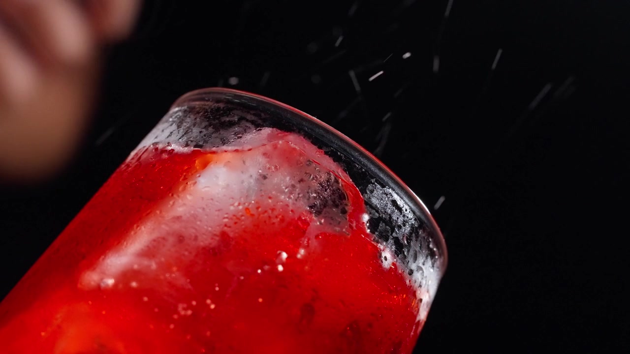 Un cocktail rosso intenso si versa in un bicchiere ghiacciato inclinato, con un flusso lento e fluido che muove un cubetto di ghiaccio.