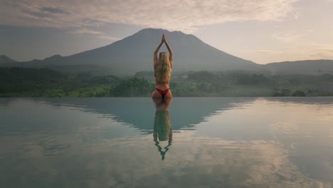 woman in bikini with hourglass figure sitting on infinity pool edge yoga salute to mount agung