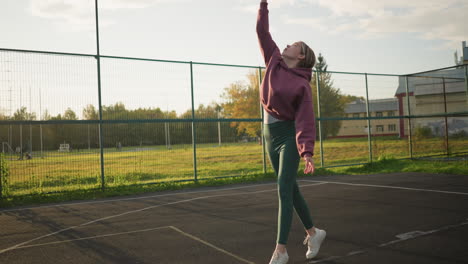 mujer atlética en leggings verdes lanza el voleibol para servir en la cancha al aire libre con campo abierto y persona caminando en el fondo, jugador enfocado preparándose para iniciar la acción del juego