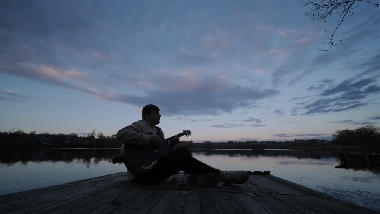 Silhouette of musician in black playing guitar sitting on pier embankment on sunset