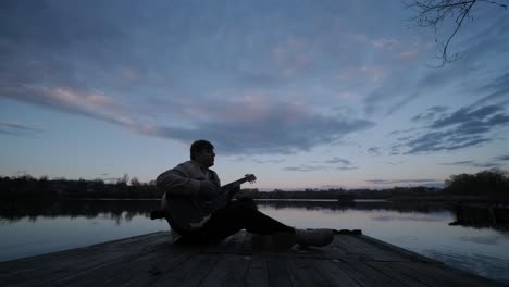 silhouette of musician in black playing guitar sitting on pier embankment on sunset