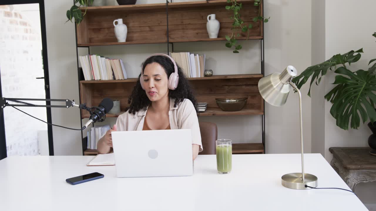 Mujer afroamericana con auriculares rosas comenzando un podcast, hablando por el micrófono y leyendo el portátil.