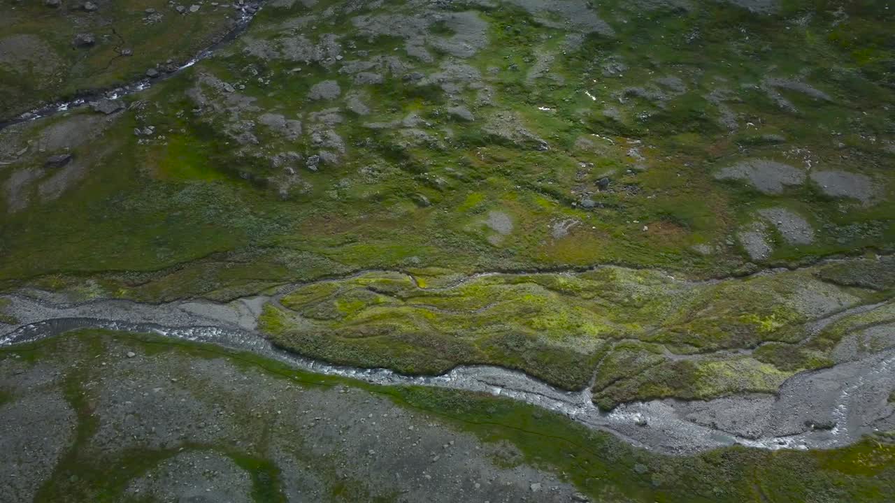 Aerial drone footage flying and gliding over a narrow river or water stream flowing on rocky granite and green moss covered Sweden landscape terrain during a cloudy day. flowing down grassy slopes.