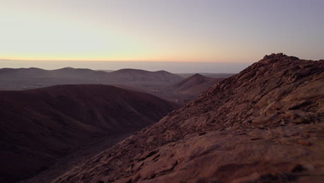 A-drone-shot-revealing-harsh,-rocky-slope-with-red-rocks-and-a-volcanic-landscape-in-the-background-during-sunset