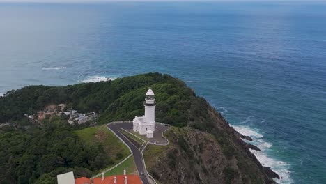 aerial view of a lighthouse perched on a green hillside with ocean waves below.