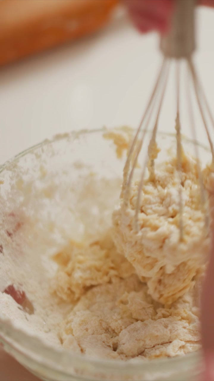 Una donna impasta in una ciotola di vetro usando una frusta, concentrandosi sulla consistenza e la densità della preparazione di pane o pasticcini. Una vista ravvicinata evidenzia la miscela che si addensa con residui di farina sui bordi.