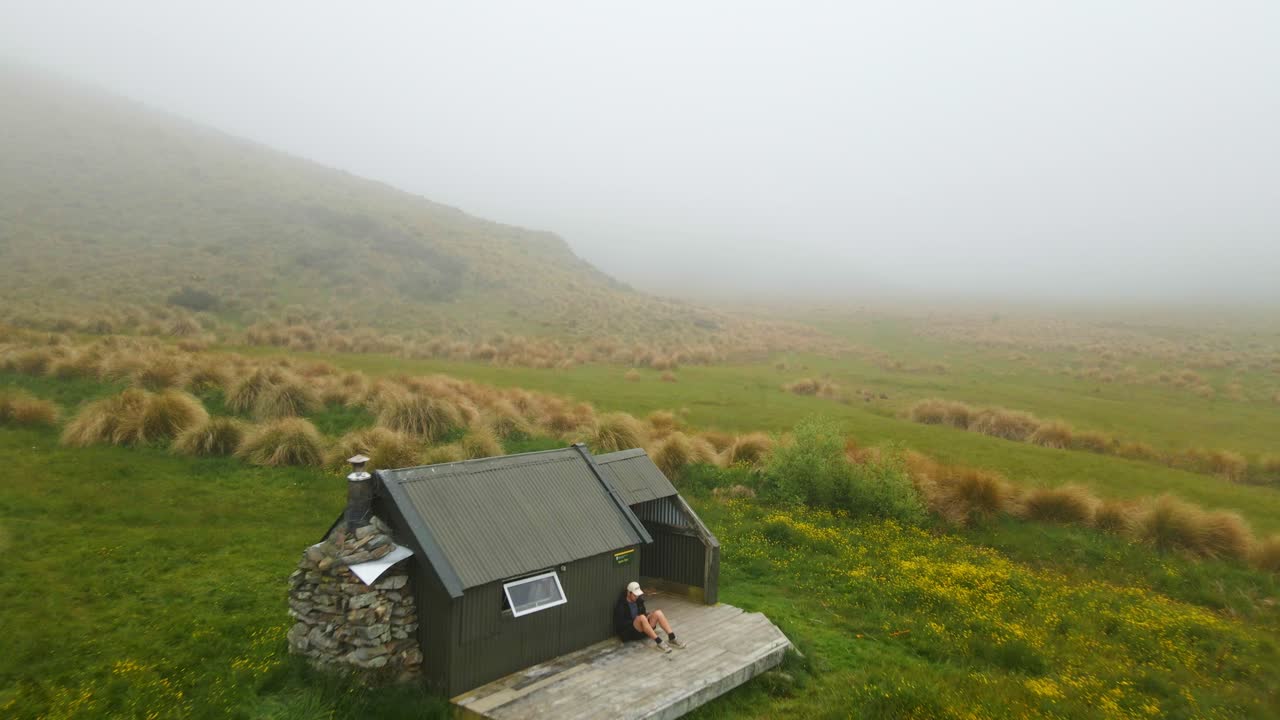 Flygende kamera avslører turgåer som sitter foran en avsidesliggende fjellhytte i tåkeforhold i Canterbury, New Zealand