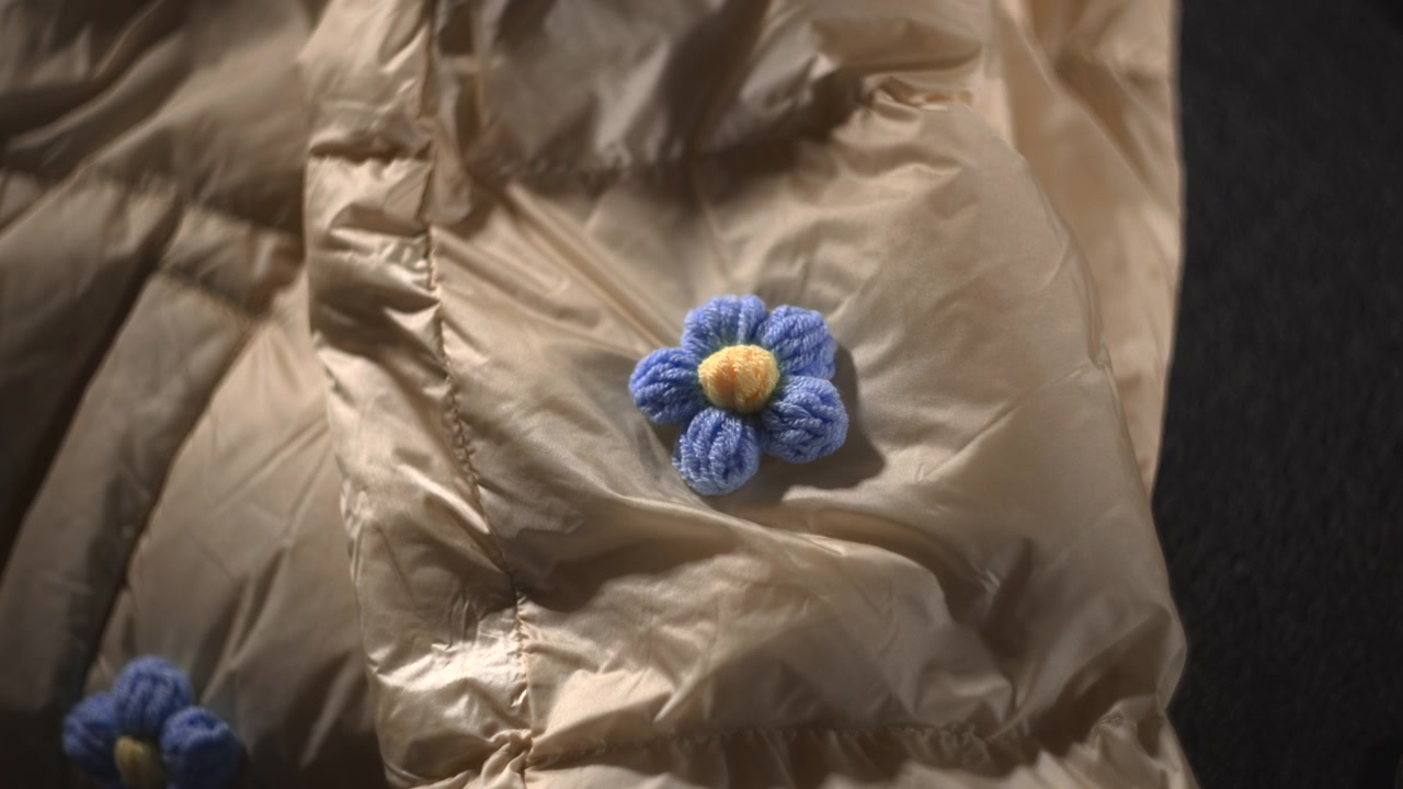 Primo piano di giacca piumino beige con fiore all'uncinetto, illuminazione morbida da studio, spilla a fiore in lana blu fatta a mano.