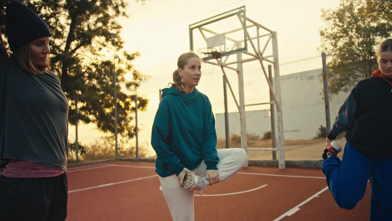 A trio of girls in sportswear warming up before playing basketball on the Red Playground fenced in by a fence near an orange basketball on a summer morning