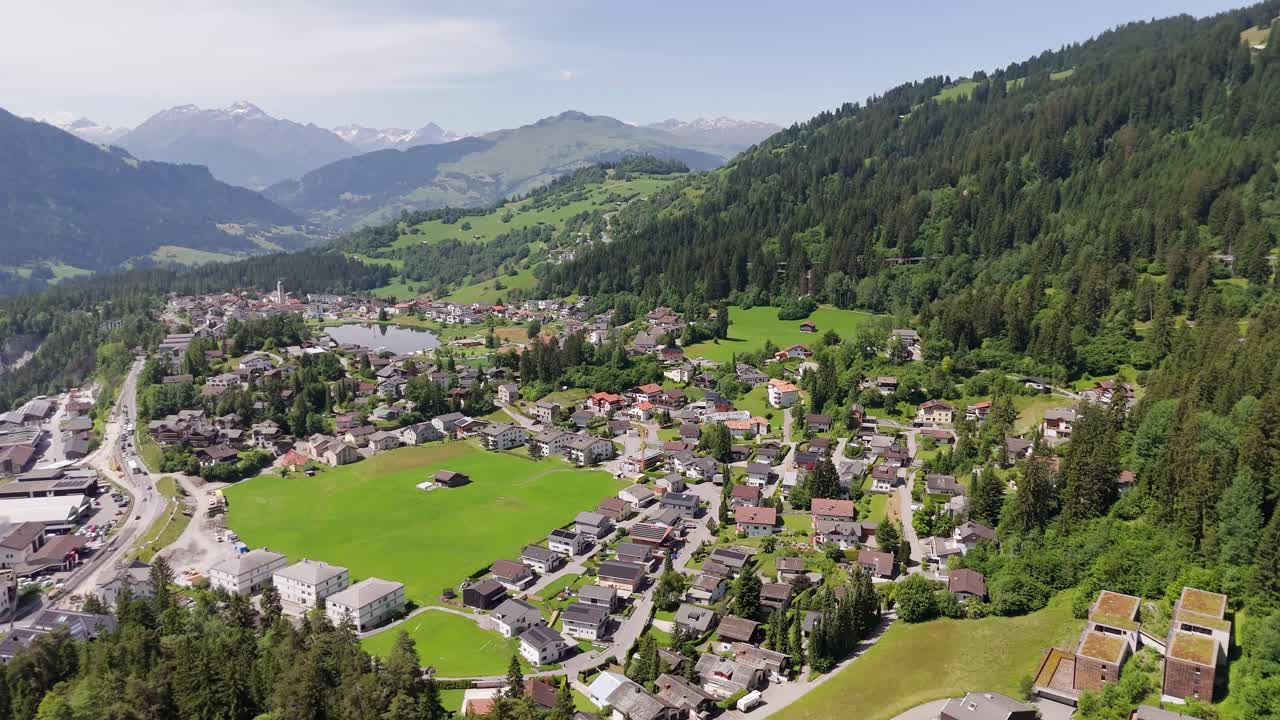 Aerial view of Swiss town with neighborhood , green pasture and cars on street. Wide shot. Sunny day in summer. Alps mountains in distance