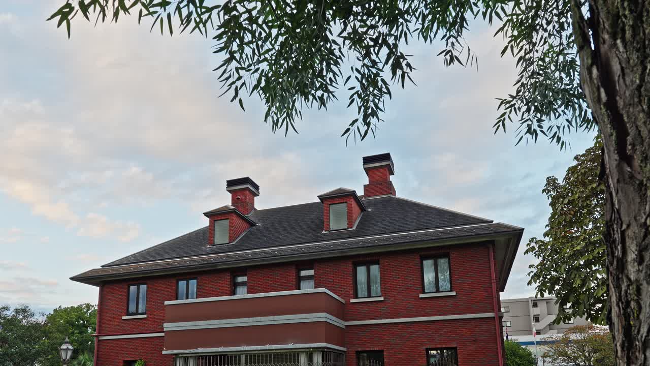Low-angle shot of a classic red brick building with chimneys, framed by weeping willow trees at dusk