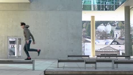 Toma-En-Cámara-Lenta-De-Un-Joven-Atleta-De-Parkour-Saltando-De-Un-Banco-A-Otro-En-Un-Lugar-Urbano