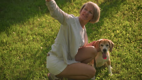 pet lover taking selfie with her dog distracted by something else outdoors in lush green garden under sunny weather, background features trees and building