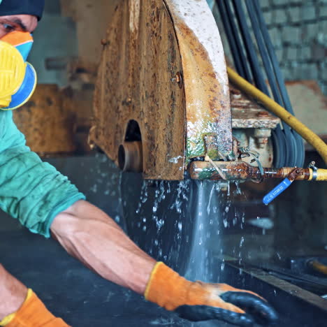 worker in uniform with protective mask puts the big stone slab into the cutting machine. circular saw in counterlight cutting stone with water for cooling.