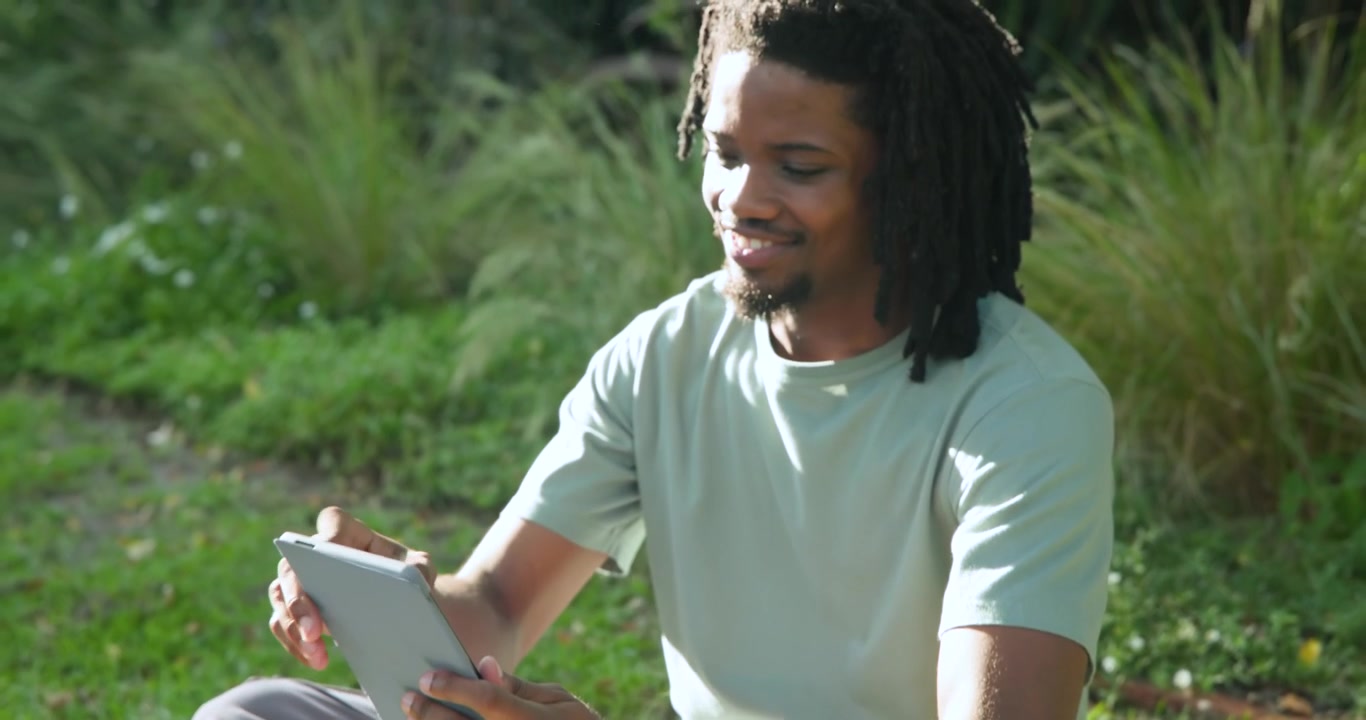 Male sitting on grass adjusting solar panel connector while using tablet to keep battery charging