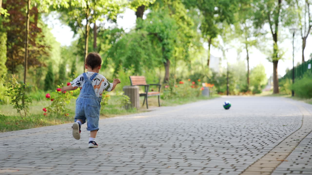 Happy Caucasian toddler runs to catch up his ball. Baby boy plays in the park in summer. Low angle view.