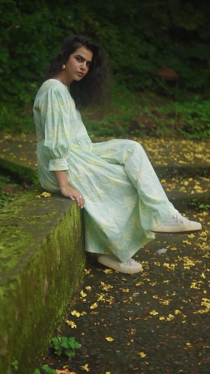 Woman in light dress and sneakers sits on a mossy stone ledge surrounded by yellow flower petals