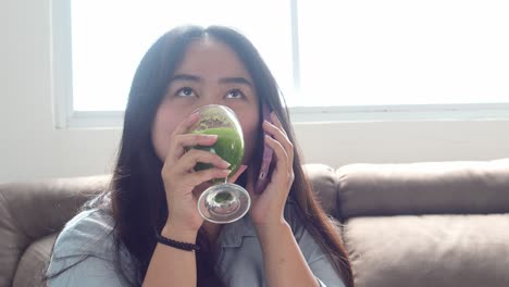 young smiling asian woman talking on the phone while drinking detox smoothie in the morning at home