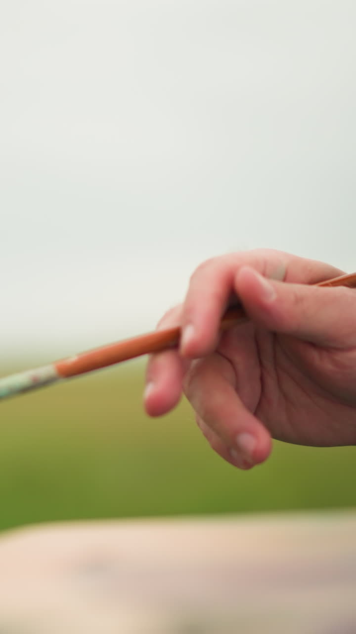 A close-up shot of an artist's hand delicately holding a paintbrush, with a blurred palette in the background. The serene, natural setting adds to the creative atmosphere