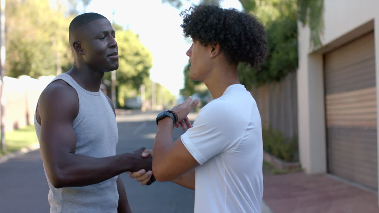 Two multiracial male friends shaking hands and talking on suburban street, enjoying hangout