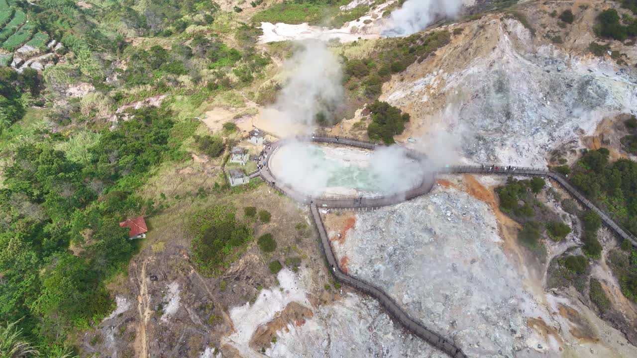 Drone scenery of Sikidang geothermal crater in Dieng, Indonesia, with visible smoke, rocky terrain, and visitors exploring the natural hot steam area