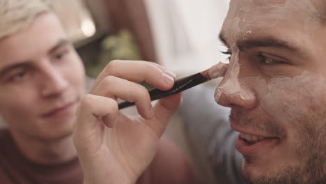 young man applying facial mask on boyfriend face