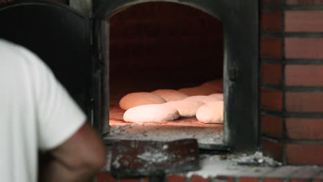 a male baker in white shirt placing the bread dough inside a traditional stone oven - making bread in bakery - medium shot