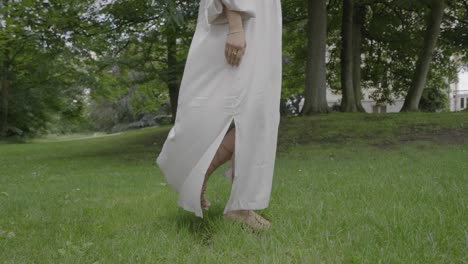 Low-angle-shot-of-a-pregnant-young-woman-wearing-a-beautiful-white-dress,-walking-through-her-lush-green-garden,-4K-120FPS
