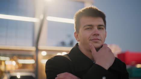 close up of investor standing outdoors in front of shopping complex, touching chin with subtle smile, mall glass reflecting golden light and structure, exuding confidence and success